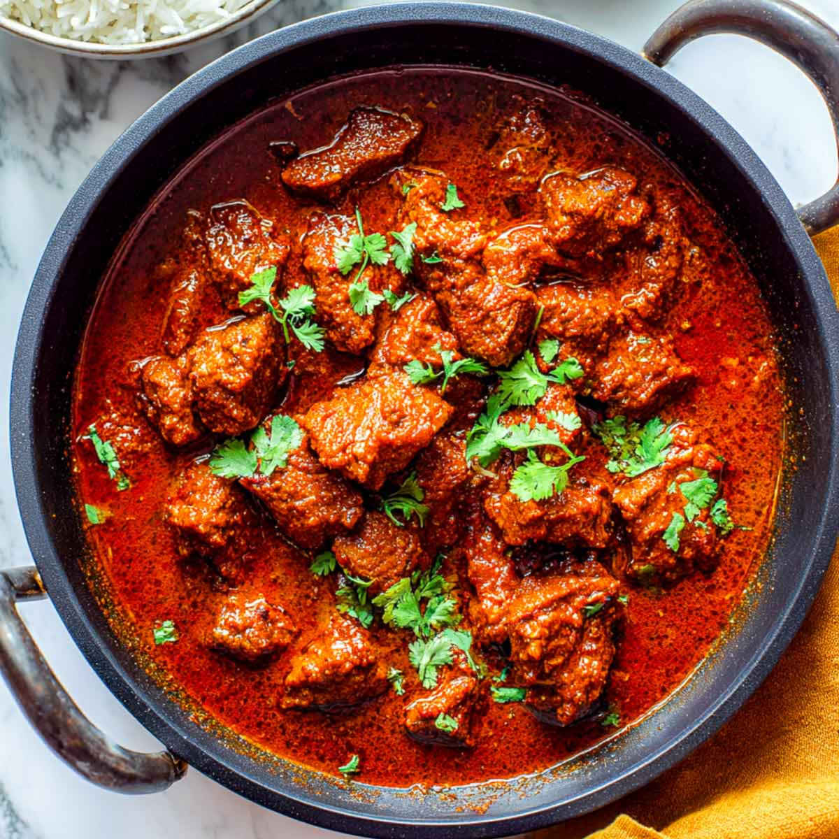 This top-down photo shows a pan of Lamb Madras simmering in a deep red gravy with tender lamb pieces and a warm, oily sheen across the surface.