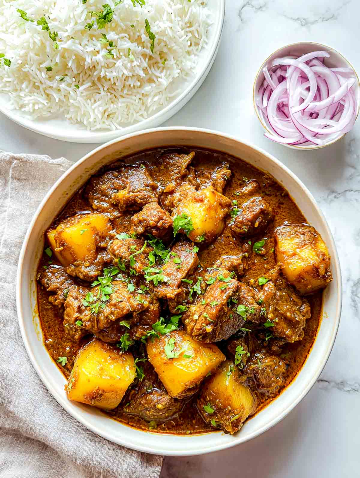 A bowl of lamb and potato curry served with rice and sliced onions.