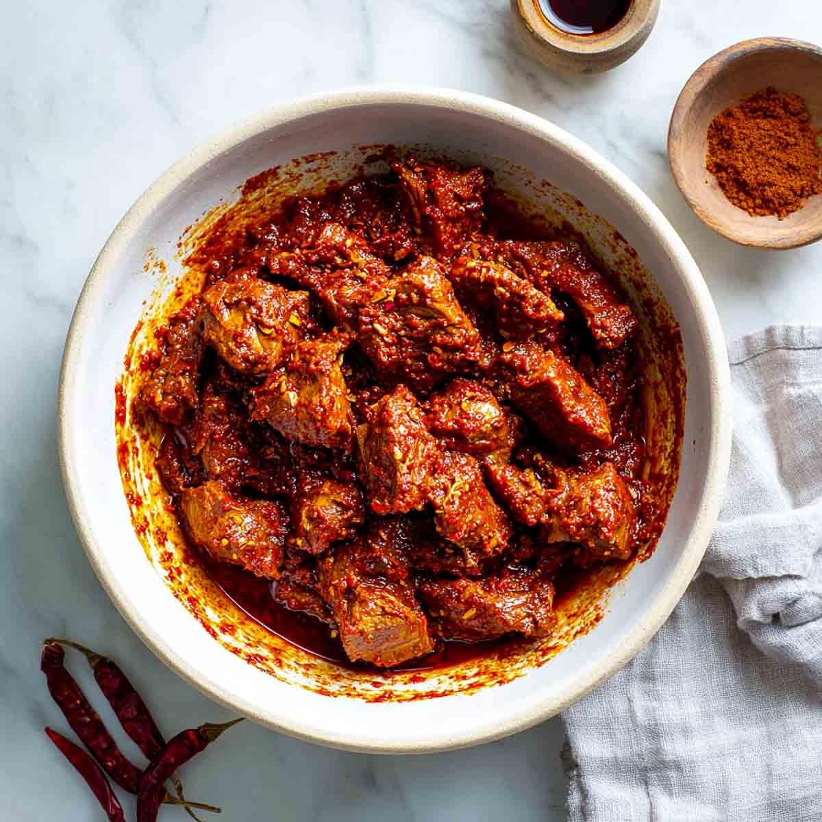 Raw lamb pieces mixed with thick vindaloo marinade in a ceramic bowl.