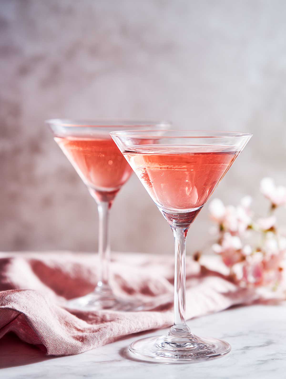 Side view of two lychee rose martinis in martini glasses on a white marble surface.