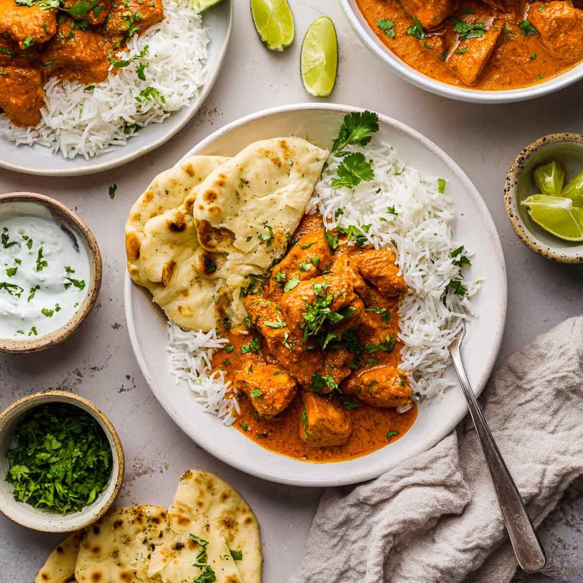 Overhead view of a plate of butter chicken with basmati rice and naan, surrounded by lime wedges, cilantro, and yogurt sauce.