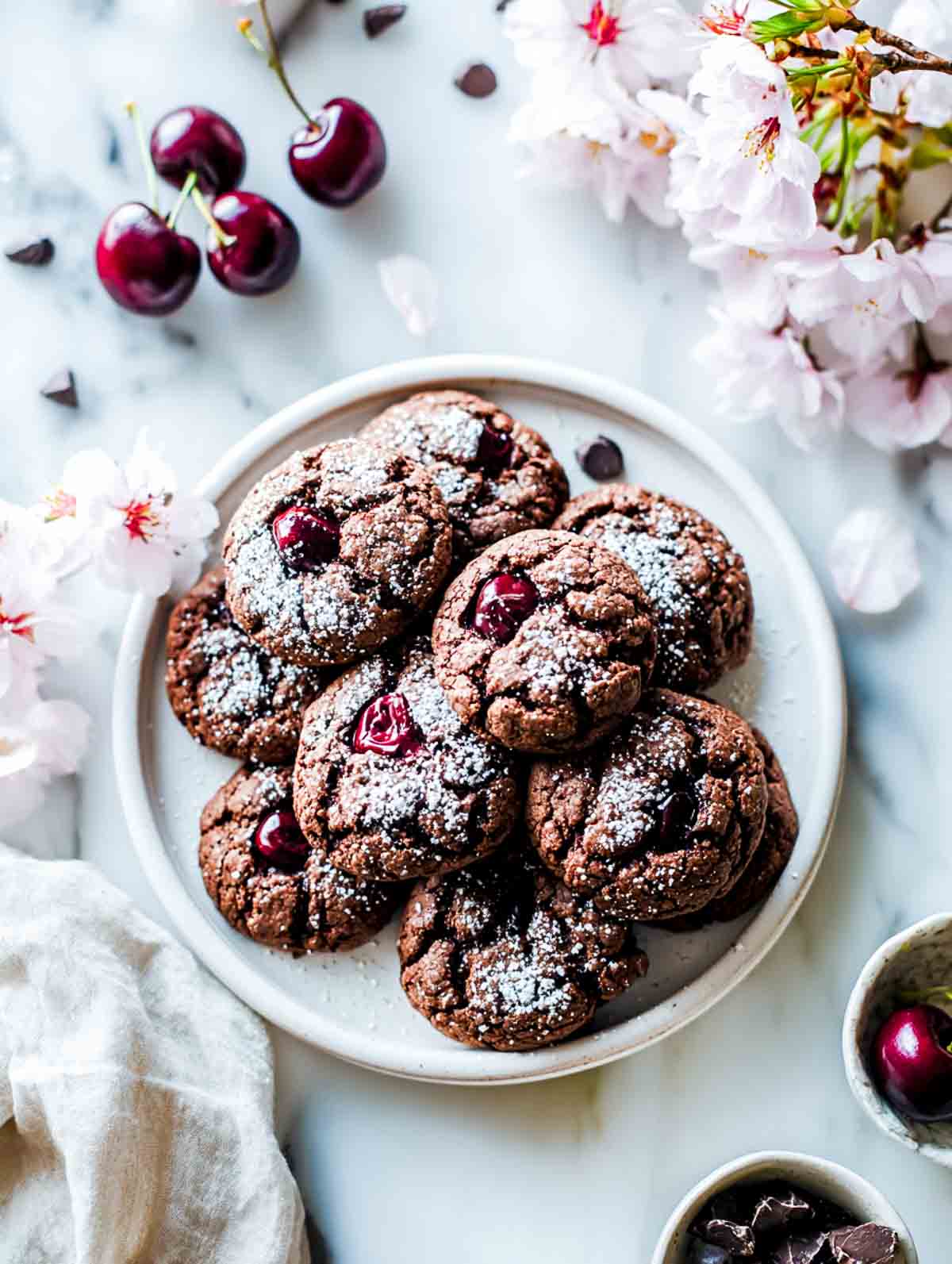 Chocolate cherry cookies on a white plate with fresh cherries and soft pink flowers.