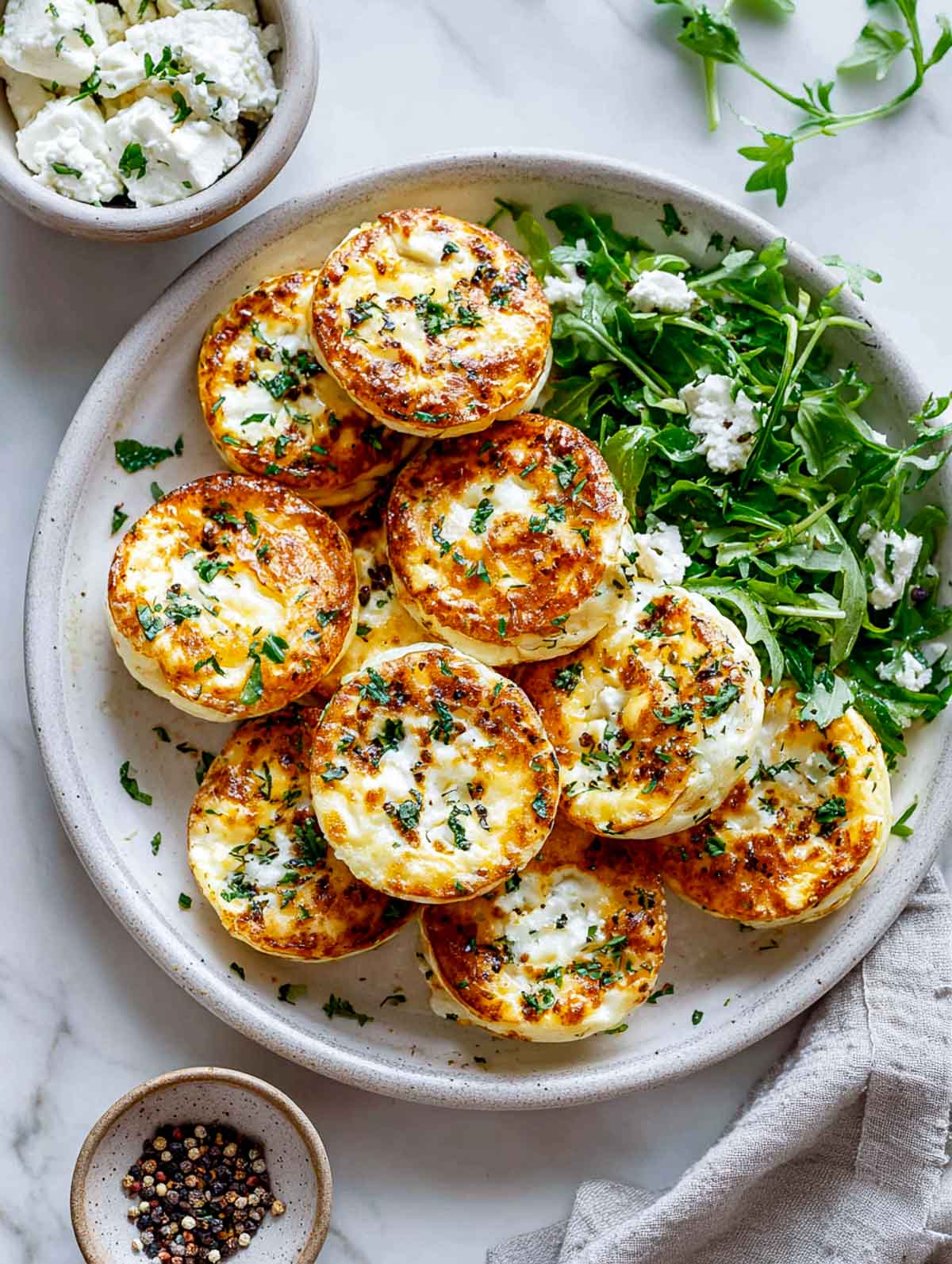 Top-down view of golden savory cottage cheese egg patties stacked on a ceramic plate with a simple arugula salad on the side, garnished with herbs and crumbled cheese, set on a bright white marble surface with small bowls of cottage cheese and black pepper nearby.