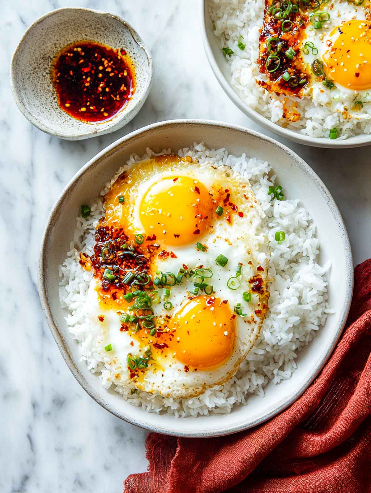 Top-down view of crispy fried eggs with jammy yolks served over garlic rice in a shallow ceramic bowl, topped with chili oil crisp and sliced green onions, with a small bowl of extra chili oil on a white marble surface.