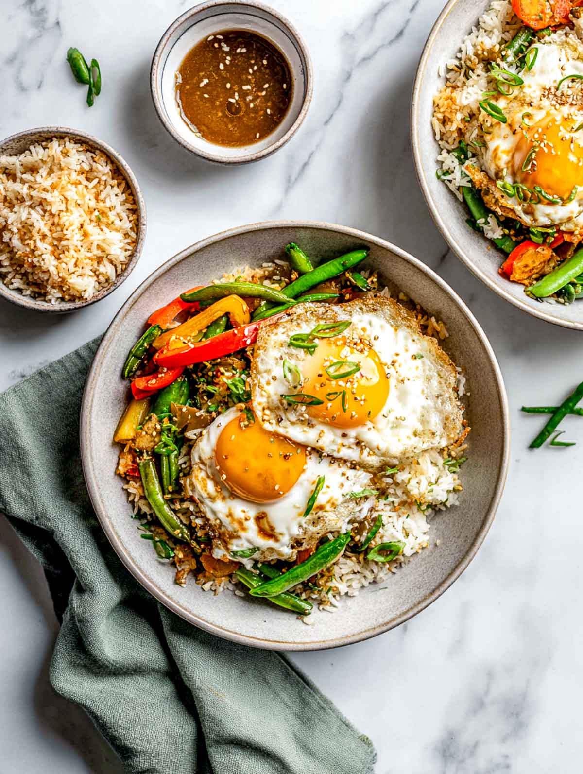 Top-down view of a crispy rice bowl topped with fried eggs and sautéed vegetables, served in a ceramic bowl on a white marble surface, with golden crunchy rice, bright jammy egg yolks, green beans and bell peppers, and small bowls of sauce nearby.