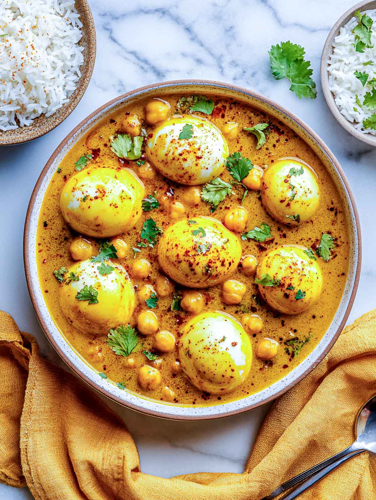 Top-down view of hard-boiled eggs simmered in a golden coconut curry with chickpeas, served in a ceramic bowl on a white marble surface, garnished with cilantro, with rice bowls and a mustard yellow napkin nearby.