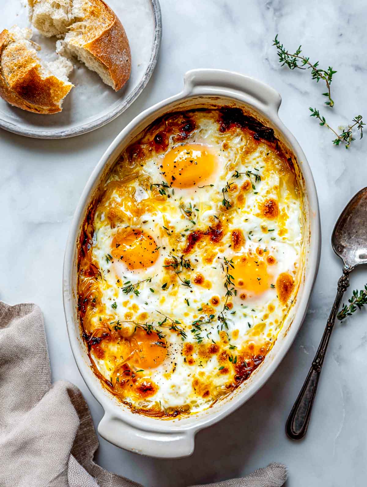 Top-down view of French onion style baked eggs in a small oval ceramic baking dish on a white marble surface, with deeply caramelized onions, softly set eggs with jammy yolks, melted golden cheese on top, fresh thyme scattered over the dish, and torn crusty bread on a plate nearby.