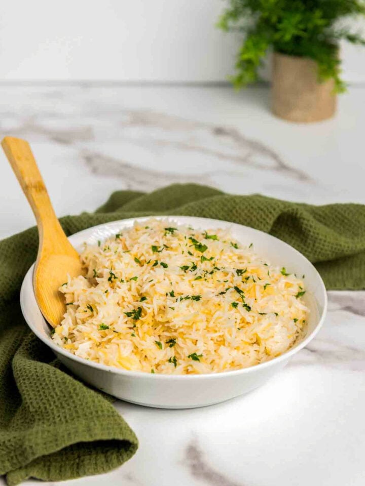 Bowl of fluffy garlic rice made with basmati and toasted vermicelli, garnished with chopped parsley, with a wooden spoon resting inside, styled on a light marble surface with a green cloth in the background.