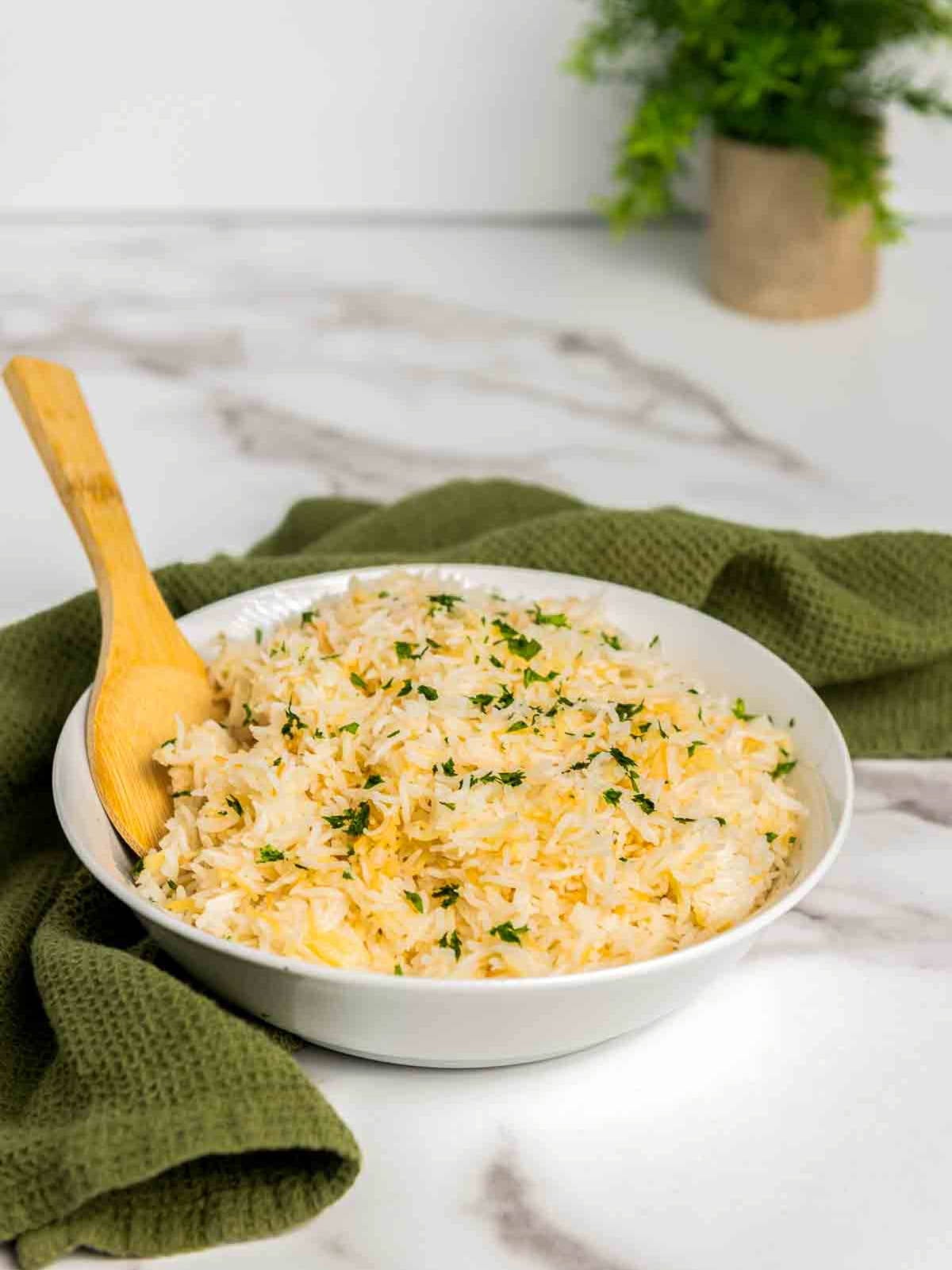 Bowl of fluffy garlic rice made with basmati and toasted vermicelli, garnished with chopped parsley, with a wooden spoon resting inside, styled on a light marble surface with a green cloth in the background.