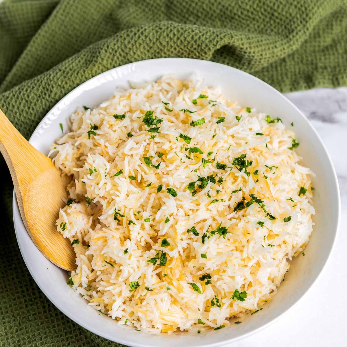 Close-up of garlic rice made with basmati and toasted vermicelli, lightly garnished with chopped parsley, served in a white bowl with a wooden spoon on a textured green cloth.