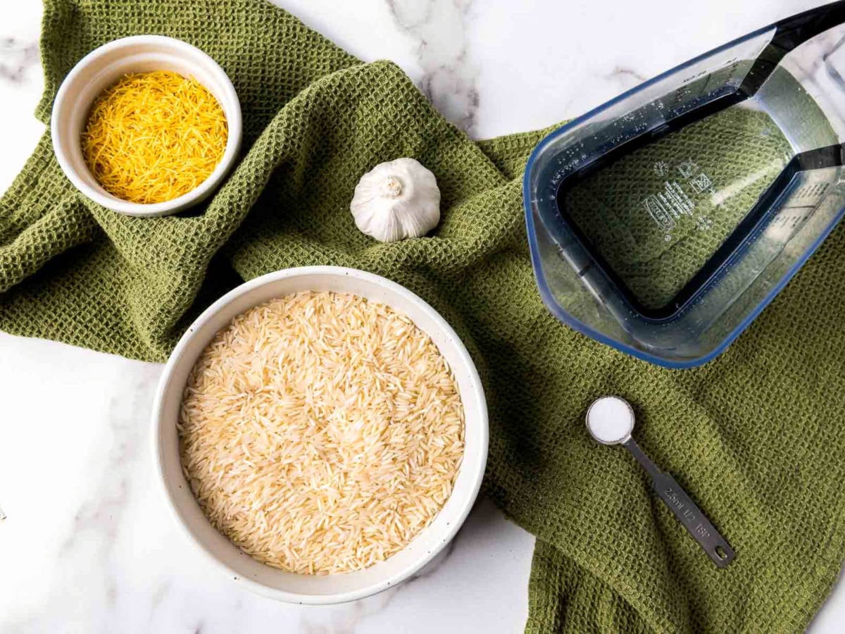Overhead view of garlic rice ingredients including uncooked basmati rice, toasted vermicelli, a whole garlic bulb, salt, and water in a measuring jug, arranged on a green textured cloth.