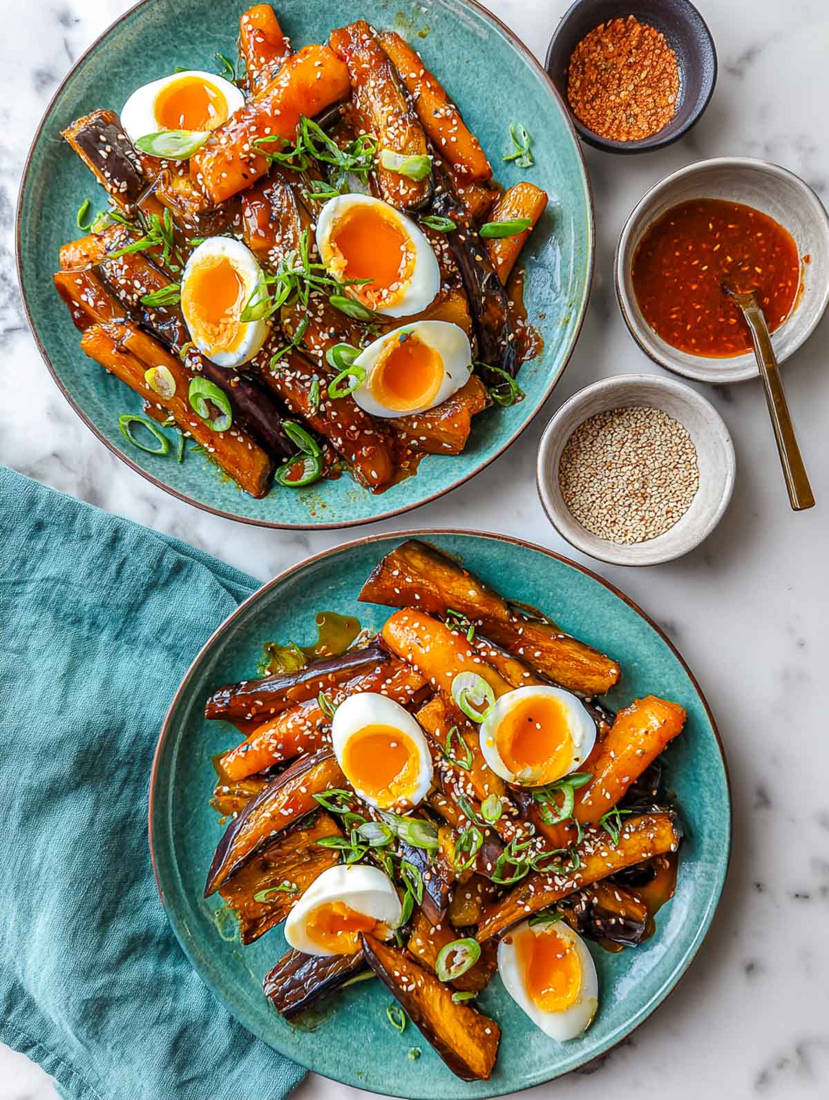 Two ceramic plates filled with roasted eggplant wedges coated in glossy gochujang sauce, topped with several soft-boiled egg halves with jammy yolks, garnished with sliced scallions and sesame seeds, set on a lightly veined white marble surface with small bowls of sauce and seeds nearby.