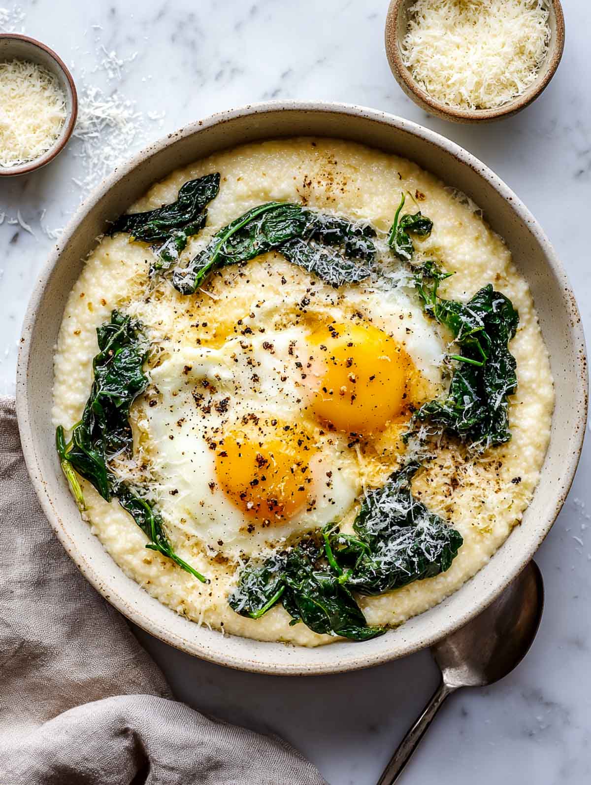 Creamy cheesy grits topped with two fried eggs, sautéed leafy greens, black pepper, and grated cheese in a ceramic bowl on a white marble surface.