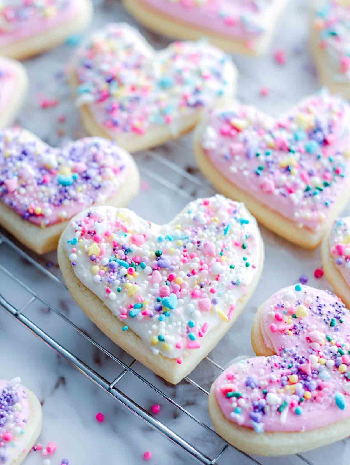 Heart-shaped sugar cookies with white and pink icing and pastel sprinkles on a cooling rack.