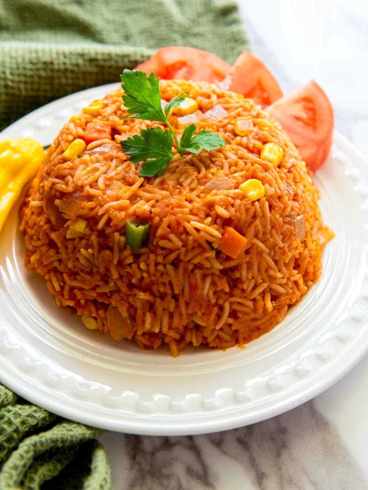 Close-up of a molded serving of jollof rice with mixed vegetables, garnished with parsley and served on a white plate with sliced tomatoes on the side.