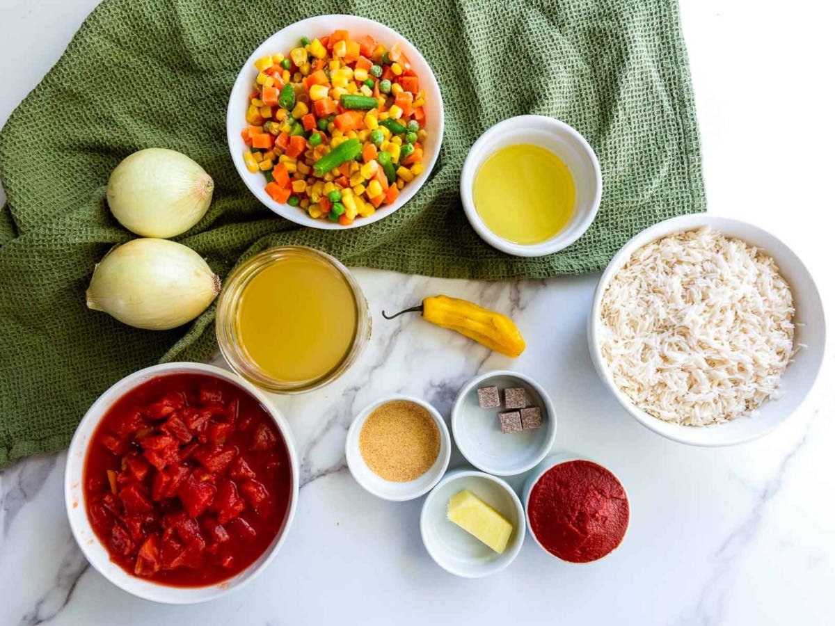 Overhead view of jollof rice ingredients arranged on a marble surface, including rice, diced tomatoes, mixed vegetables, onions, stock, oil, tomato paste, habanero pepper, seasoning cubes, and spices.