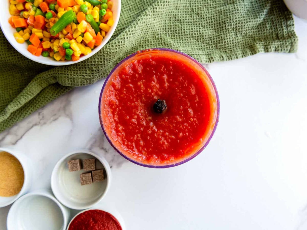 Overhead view of blended tomato mixture in a food processor bowl, with jollof rice ingredients like mixed vegetables, seasoning cubes, and tomato paste arranged nearby on a marble surface.