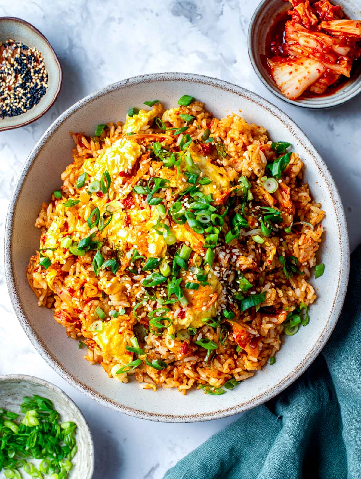Top-down view of kimchi egg fried rice in a wide ceramic bowl on a white marble surface, showing fluffy rice mixed with chopped kimchi, soft scrambled eggs folded through, sesame seeds, and sliced green onions, with small bowls of kimchi and sesame nearby.