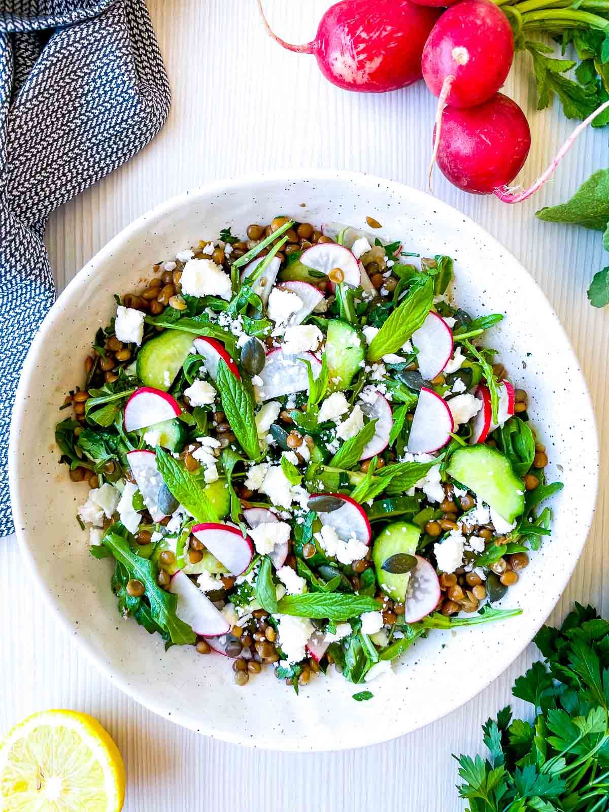 Overhead view of a fresh lentil salad with sliced radishes, cucumbers, leafy greens, mint, and crumbled cheese in a white bowl, with lemon and herbs on the side.
