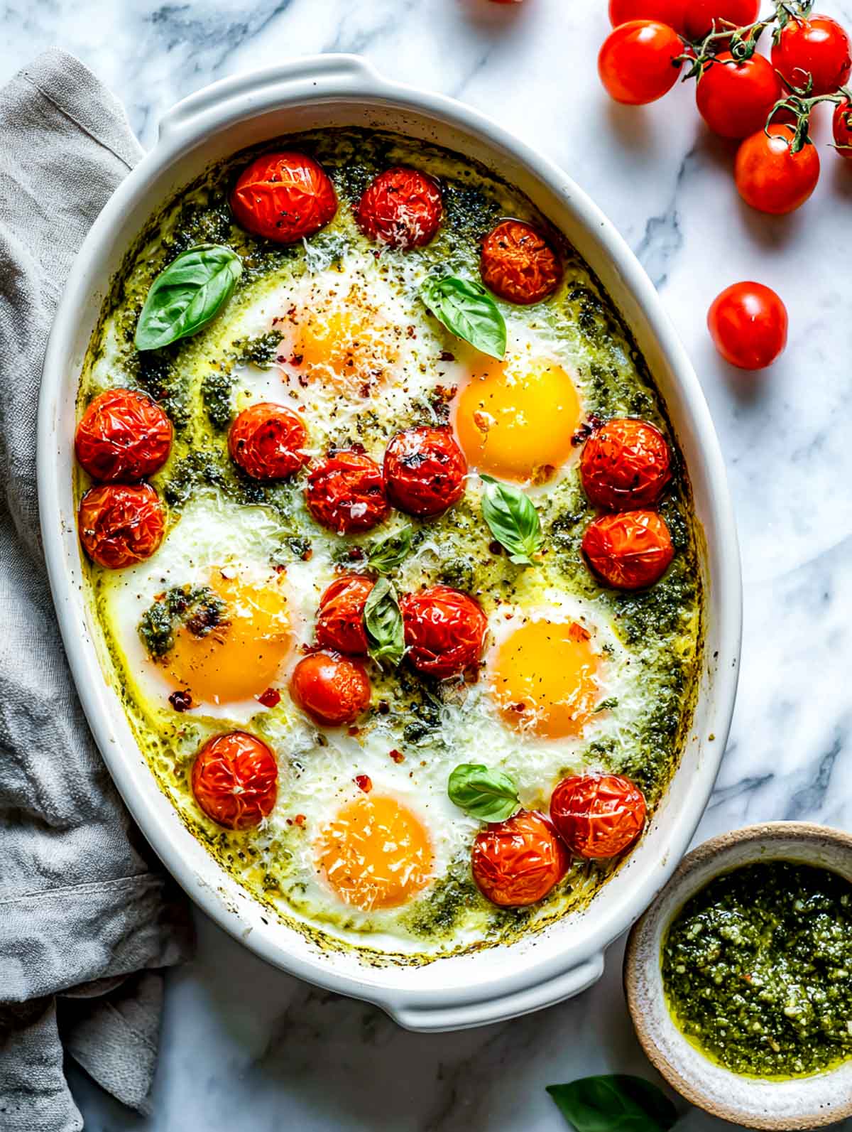 Top-down view of pesto baked eggs in a white ceramic baking dish on a white marble surface, showing eggs baked into green pesto with jammy yolks, blistered cherry tomatoes scattered throughout, fresh basil leaves on top, and small bowls of pesto and fresh tomatoes nearby.