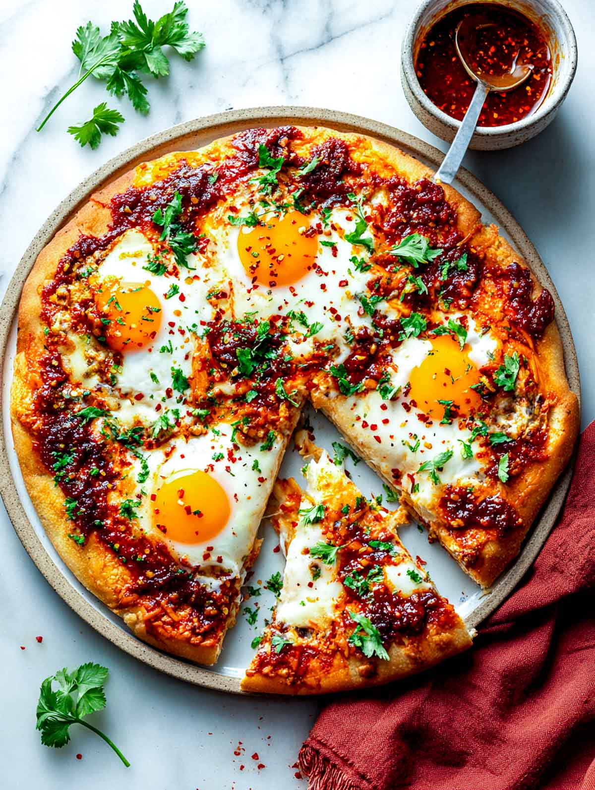 Top-down view of spicy shakshuka flatbread pizza baked on a round ceramic dish, topped with rich tomato pepper sauce, baked eggs with softly set whites and jammy yolks, chopped parsley, and red chili flakes, with one slice removed to show the interior.