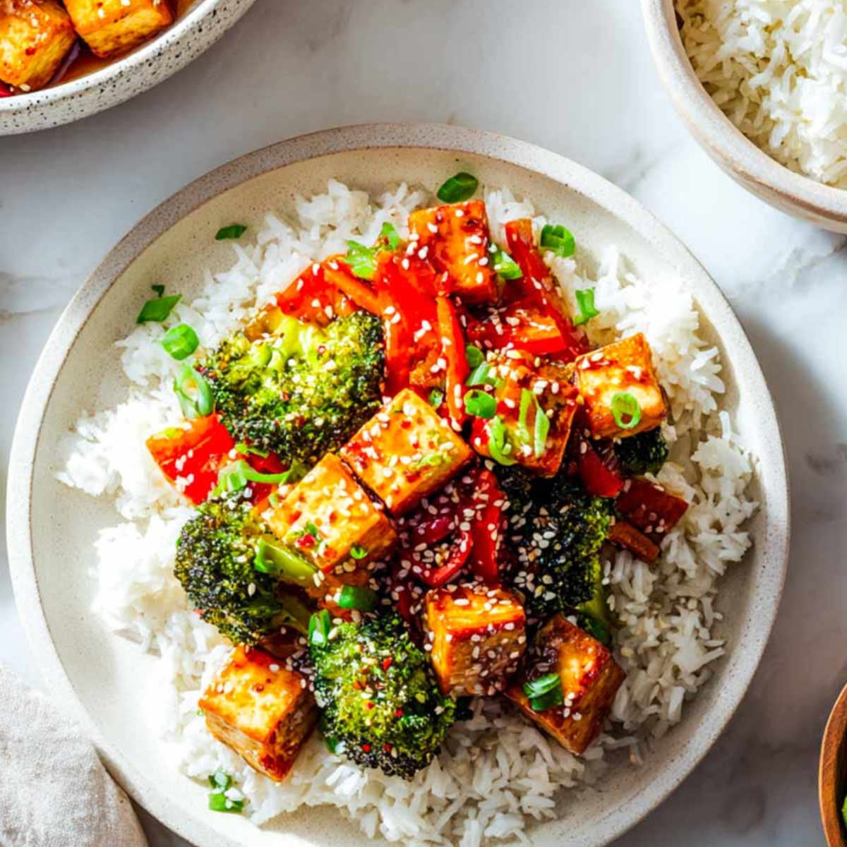 Top-down view of a single serving of tofu and broccoli over white rice in an off-white ceramic bowl, garnished with sesame seeds and green onion.