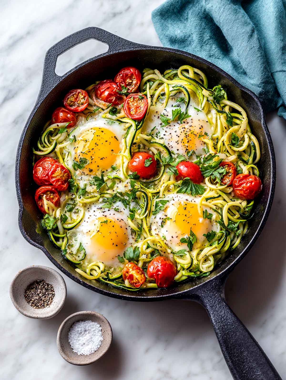 Top-down view of a cast iron skillet filled with spiralized zucchini noodles, four baked eggs with set whites and jammy yolks, blistered cherry tomatoes, fresh parsley, and cracked black pepper on a white marble surface with a muted teal napkin partially covering the skillet handle.