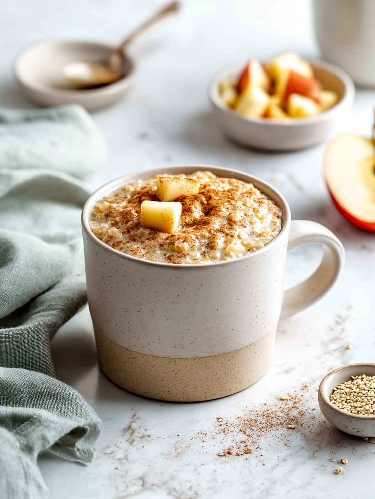 A two-tone ceramic mug filled with creamy quinoa porridge topped with diced apples and a sprinkle of cinnamon, set on a white marble surface with a sage green napkin and small bowls of apples and quinoa nearby.