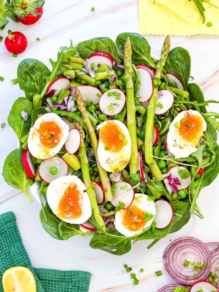 Asparagus salad with soft-boiled eggs, radishes, peas, red onion, and leafy greens on a white plate.
