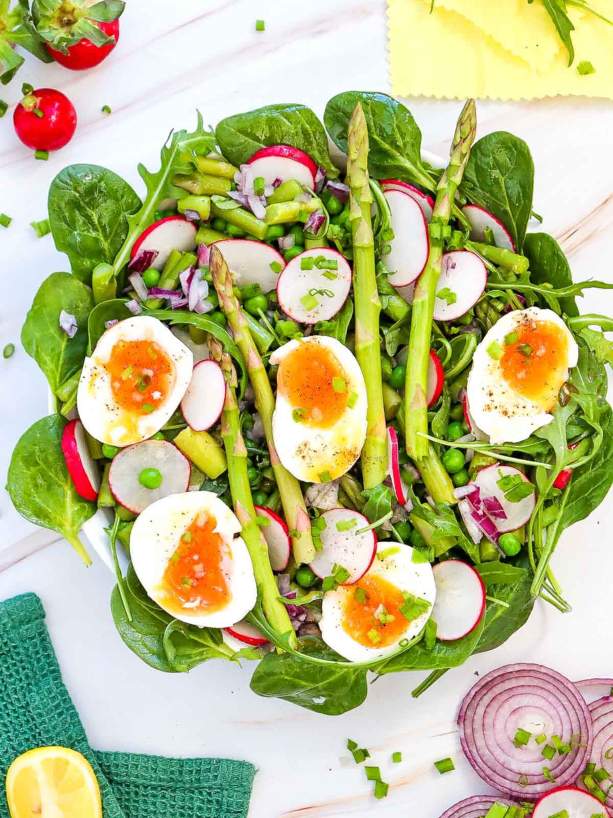 Asparagus salad with soft-boiled eggs, radishes, peas, red onion, and leafy greens on a white plate.