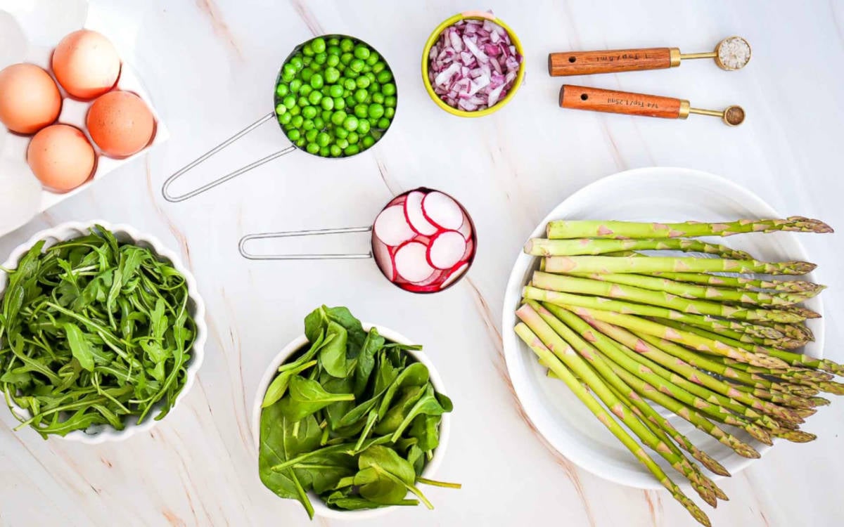 Ingredients for asparagus salad: fresh asparagus, eggs, peas, sliced radishes, red onion, arugula, and spinach on a white surface.