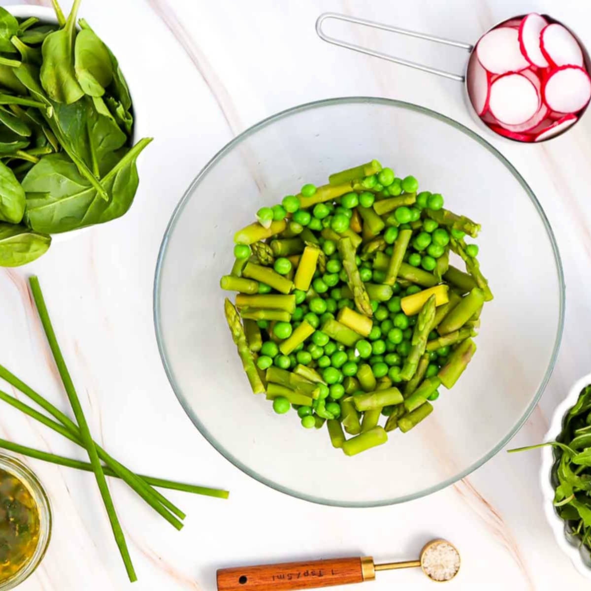 Glass bowl filled with cooked asparagus and green peas, surrounded by fresh salad ingredients.