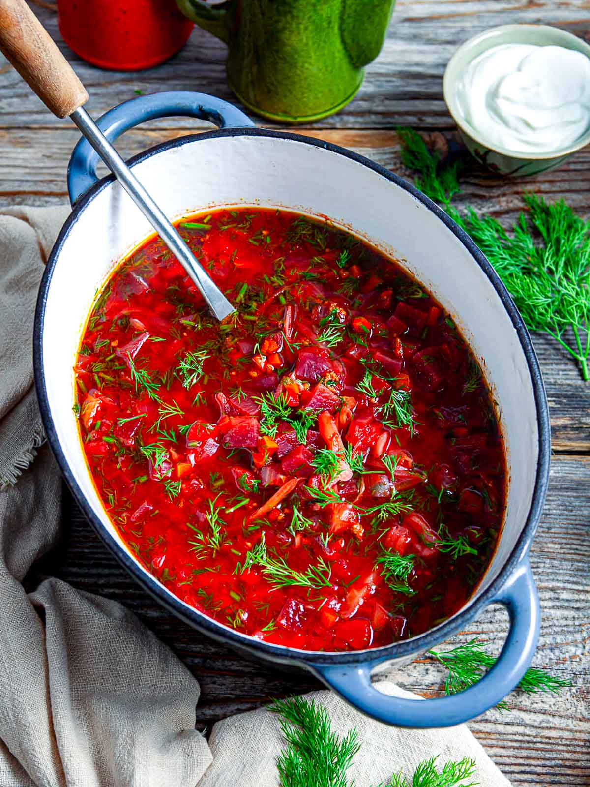 A pot of vibrant beet borscht soup garnished with fresh dill, served with a spoon on a rustic wooden table.