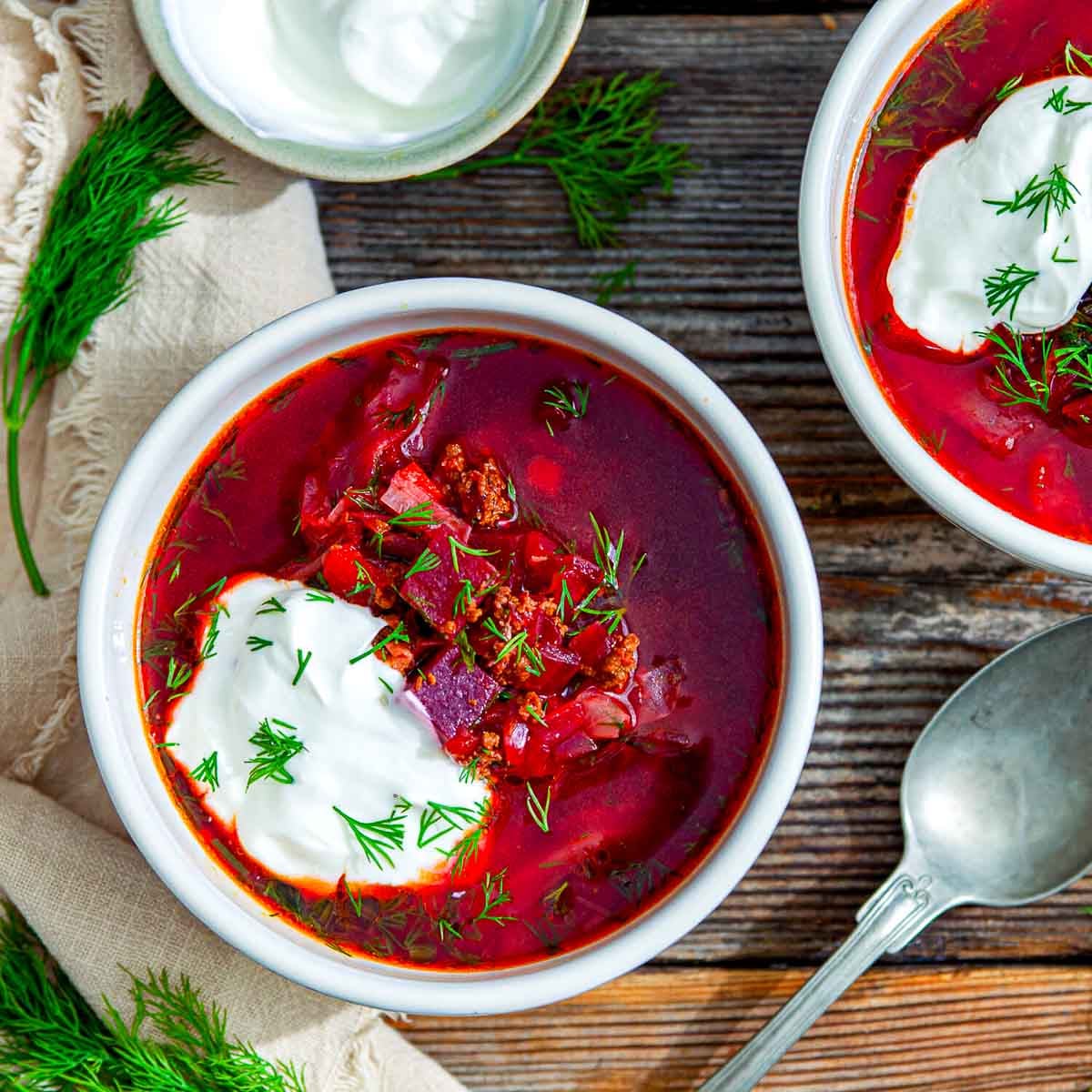 Two bowls of vibrant beet borscht served with generous dollops of sour cream and sprinkled with fresh dill, set on a rustic wooden table beside a spoon and extra herbs.
