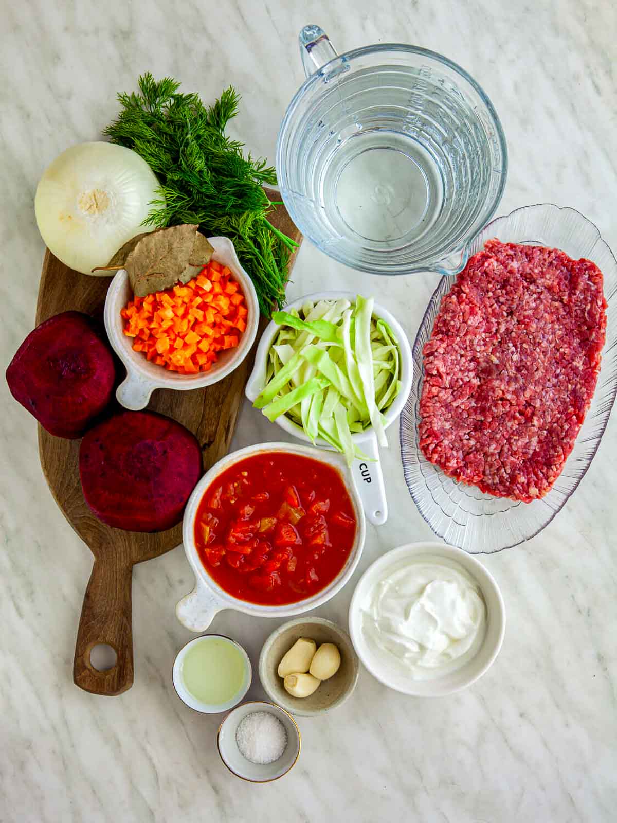 An overhead view of beet borscht ingredients arranged on a light marble surface, including fresh beets, ground beef, chopped carrots, sliced leeks, diced tomatoes, onion, garlic, dill, sour cream, and seasonings, with a measuring jug of water.