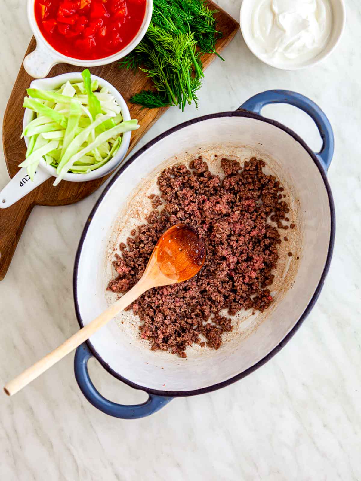 Ground beef browning in a large pot with a wooden spoon, surrounded by prepped borscht ingredients on a countertop.