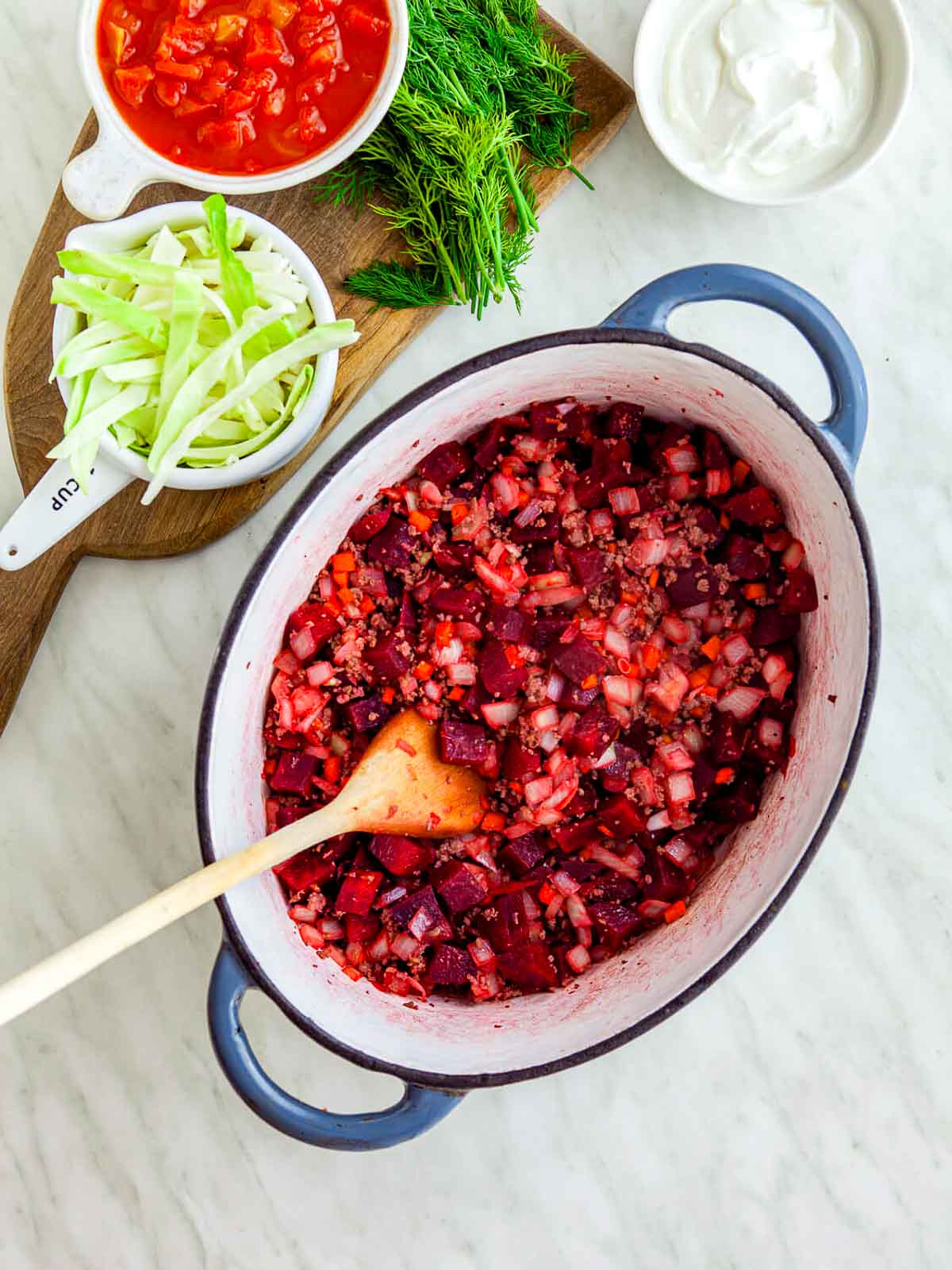 Diced beets, onions, and carrots cooking with ground beef in a large pot, with other borscht ingredients nearby.