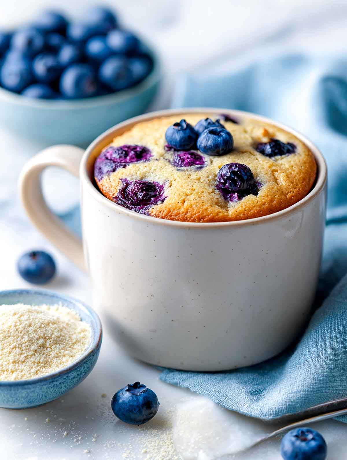 A beige ceramic mug holding a golden almond flour muffin studded with baked blueberries, set on a white marble countertop with a soft blue napkin and a small bowl of almond flour and fresh blueberries nearby.