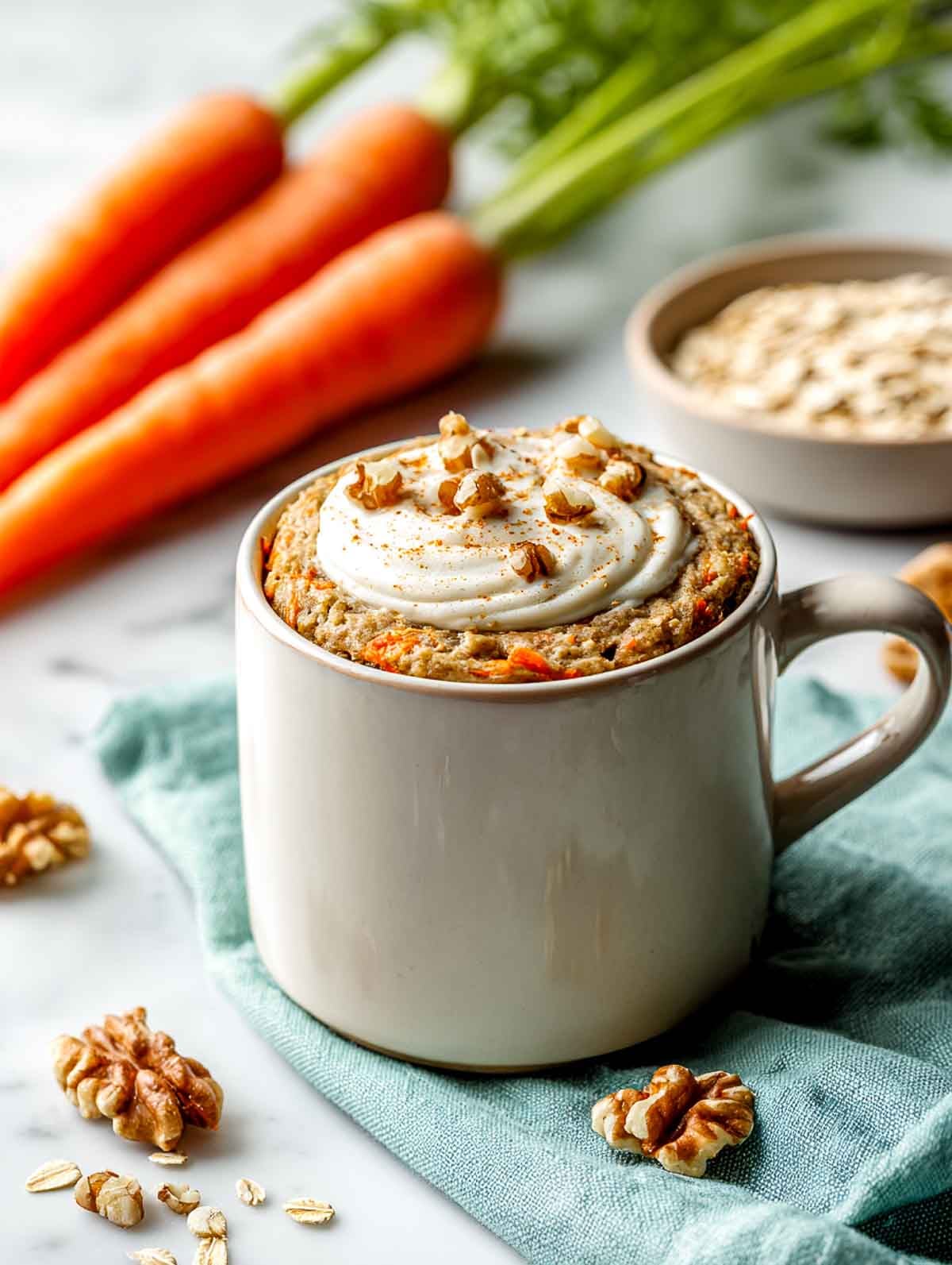 A bright angled view of an ivory ceramic mug filled with a carrot cake oatmeal protein mug bake, topped with a light Greek yogurt swirl and chopped walnuts, set on a white marble countertop with fresh carrots, a teal linen napkin, and a small bowl of oats nearby.