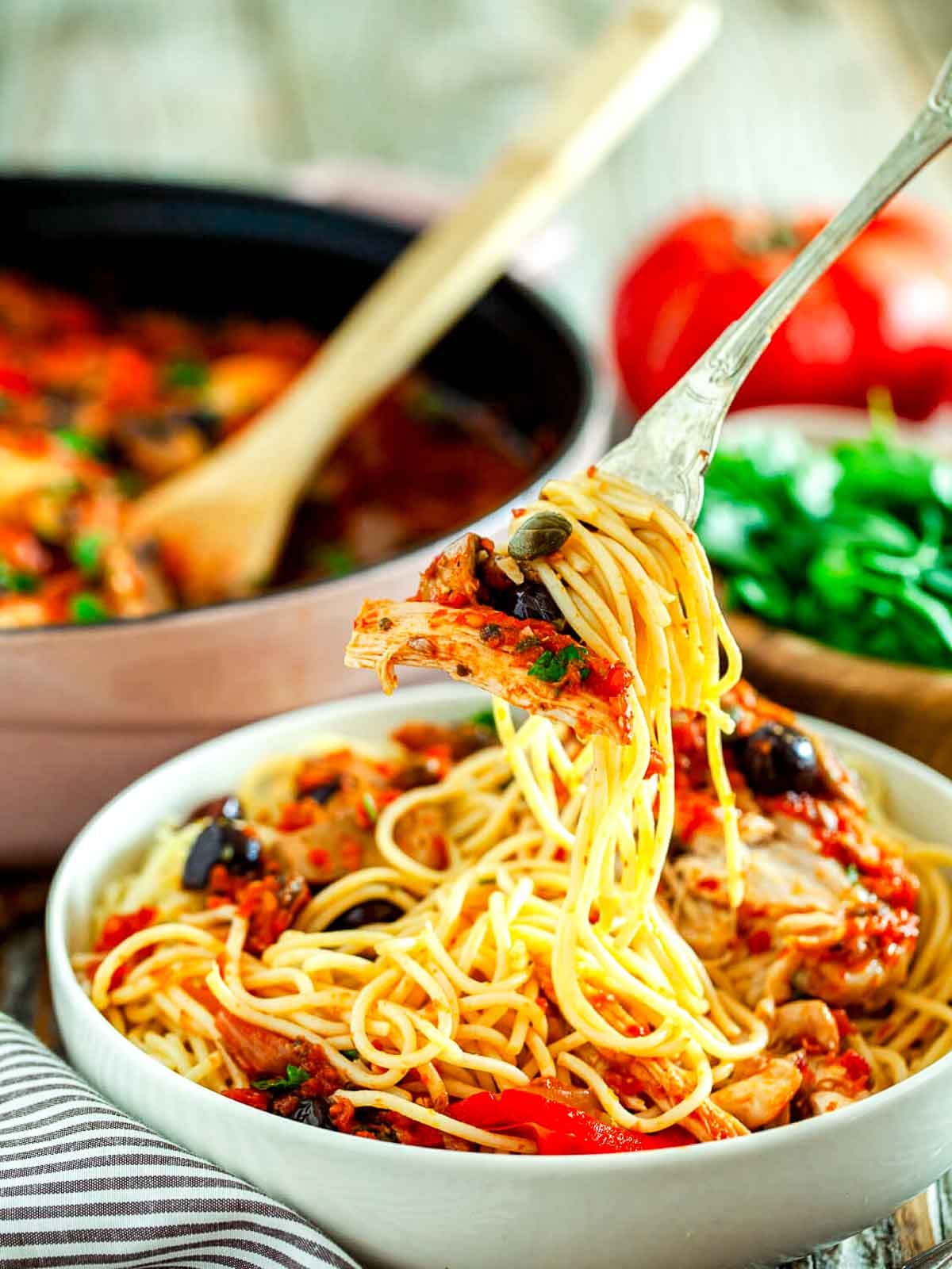 A fork lifting spaghetti and chicken cacciatore coated in rich tomato sauce, with olives and capers visible in the dish.