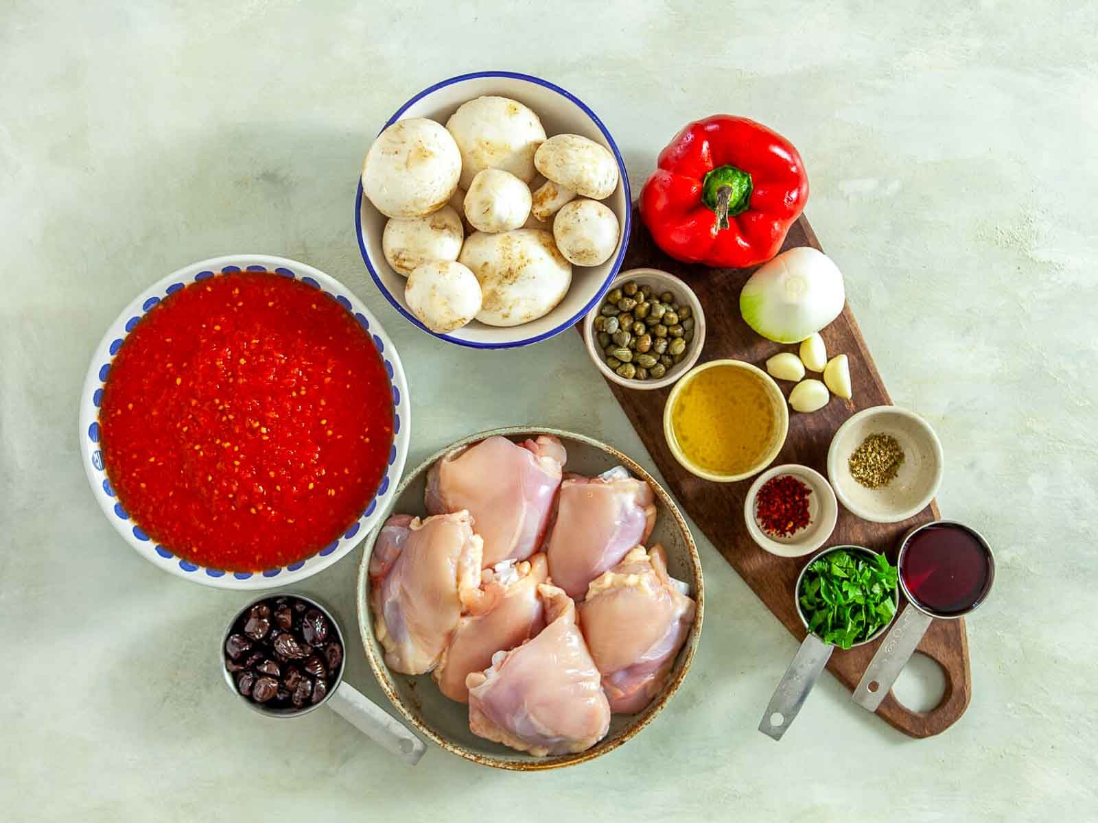 Ingredients for classic chicken cacciatore arranged on a countertop: bone-in chicken thighs, crushed tomatoes, mushrooms, red bell pepper, onion, garlic, capers, olives, olive oil, dried herbs, red pepper flakes, fresh parsley, and red wine.