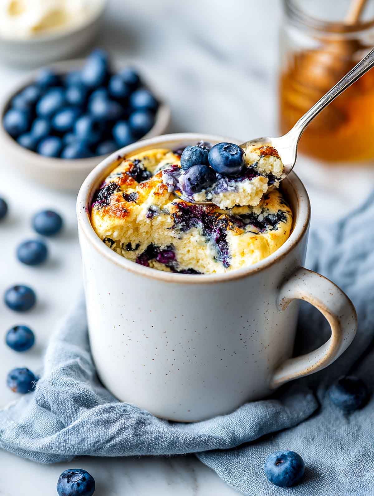 A bright, cool-toned side view of a white ceramic mug filled with a cottage cheese blueberry protein mug bake, topped with fresh blueberries and a spoon lifting a fluffy scoop, set on a white marble countertop with a soft blue linen napkin, a small bowl of blueberries, and a jar of honey nearby.