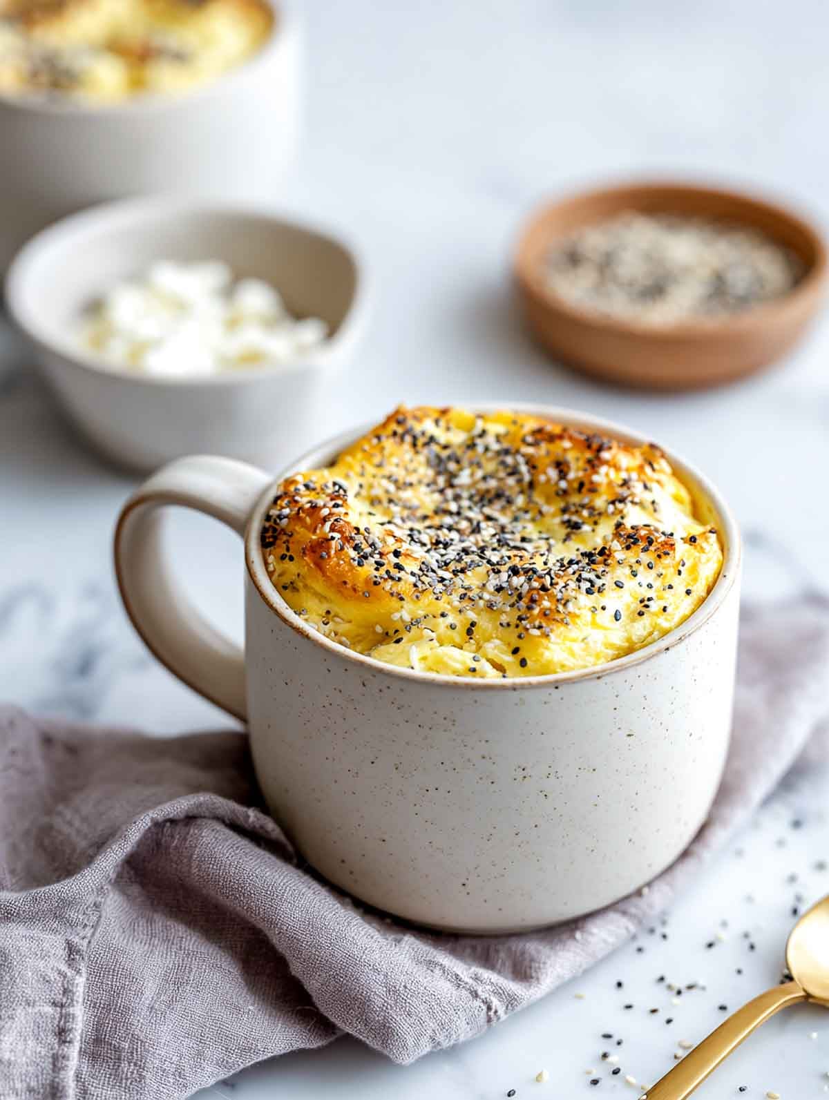 A warm cream ceramic mug filled with a lightly puffed egg bake mixed with cottage cheese and topped with everything bagel seasoning, set on a white marble countertop with a lavender napkin and small bowls of seasoning and cottage cheese nearby.