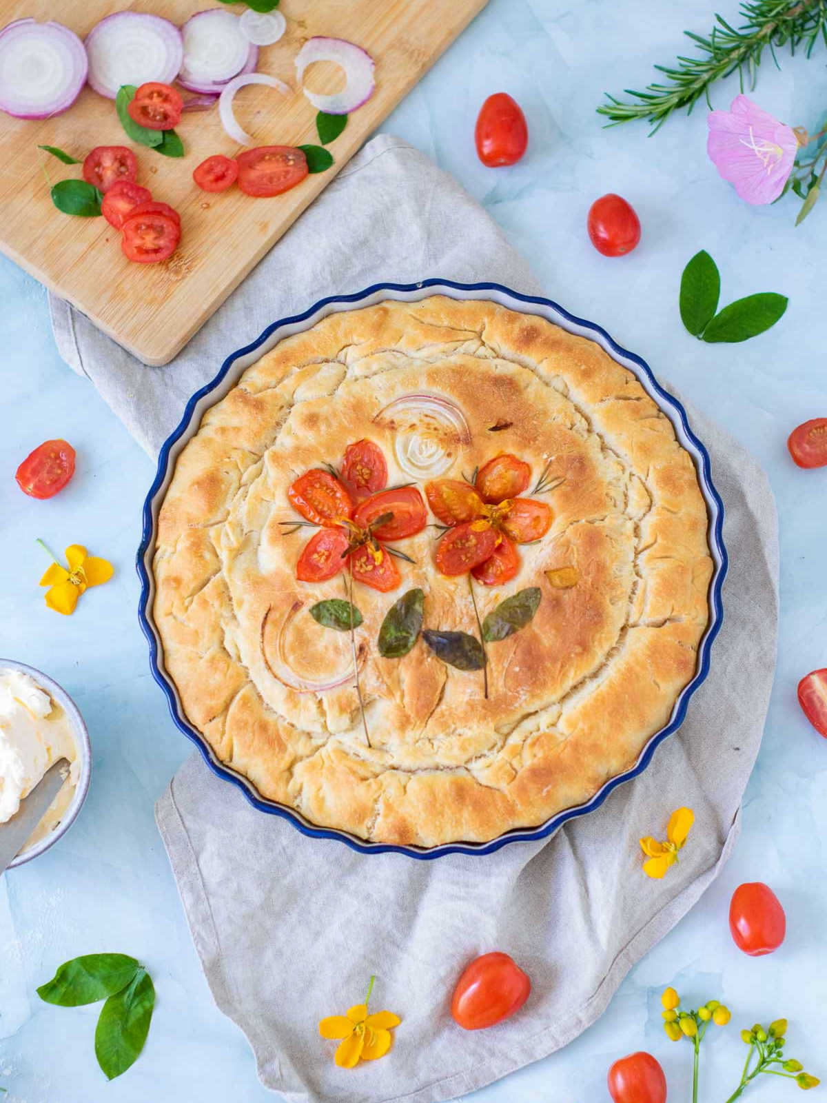 A round focaccia with cherry tomatoes on a white surface with scattered tomato and basil in the background.