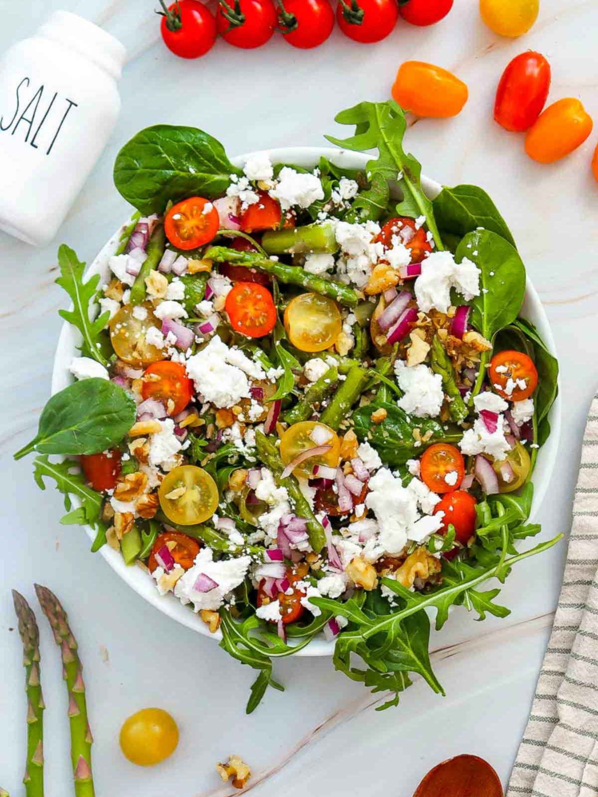 Bowl of hearty salad with arugula, asparagus, cherry tomatoes, red onion, walnuts, and crumbled goat cheese