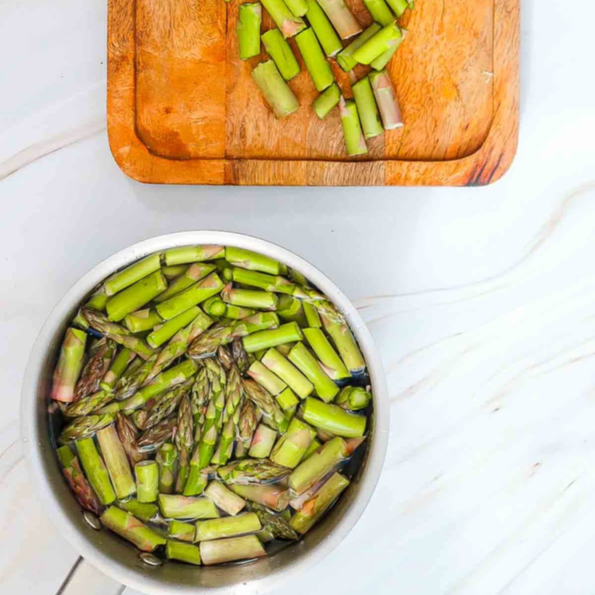Chopped asparagus soaking in water in a saucepan, with trimmed ends on a wooden cutting board