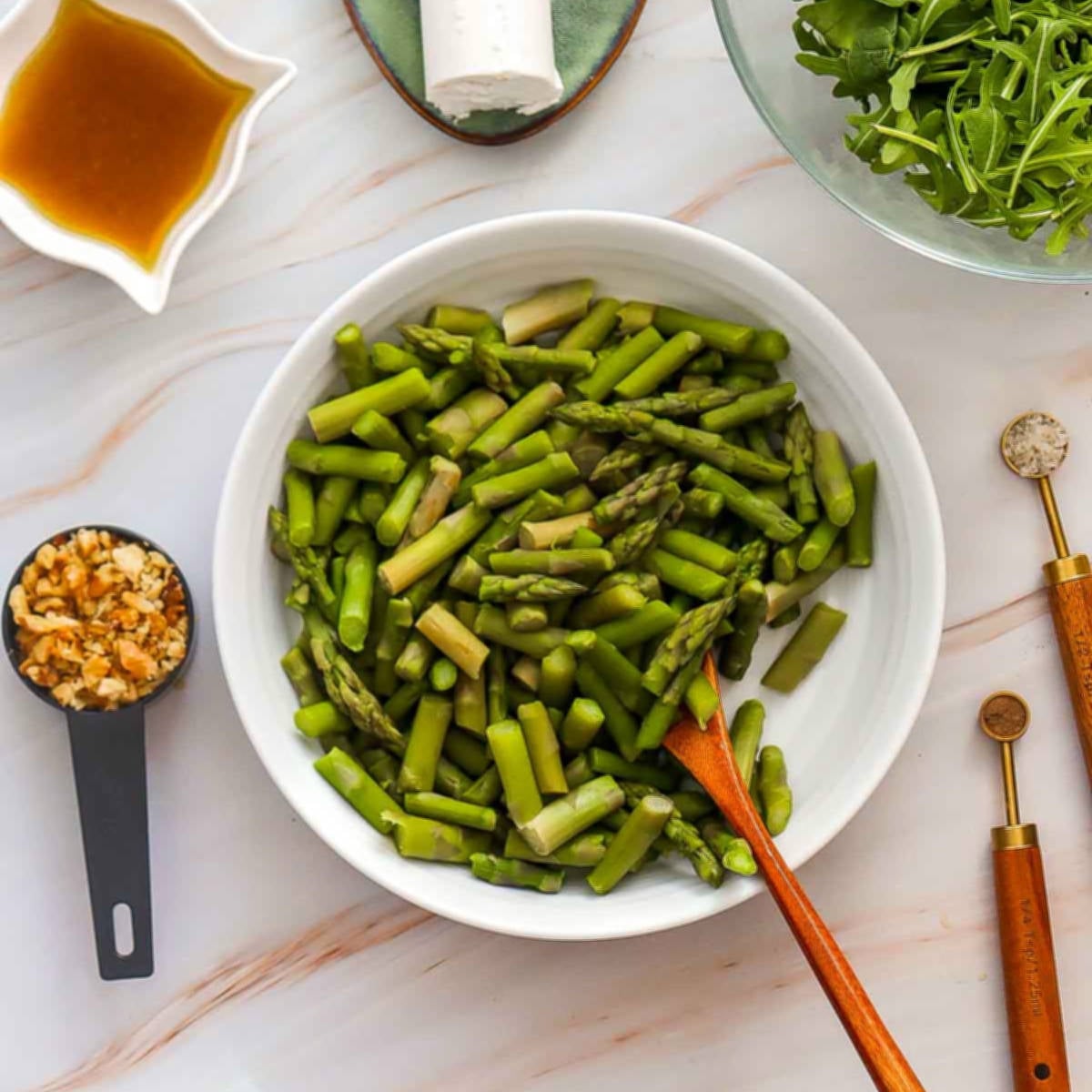 Cooked chopped asparagus in a white bowl with dressing, goat cheese, and chopped nuts nearby