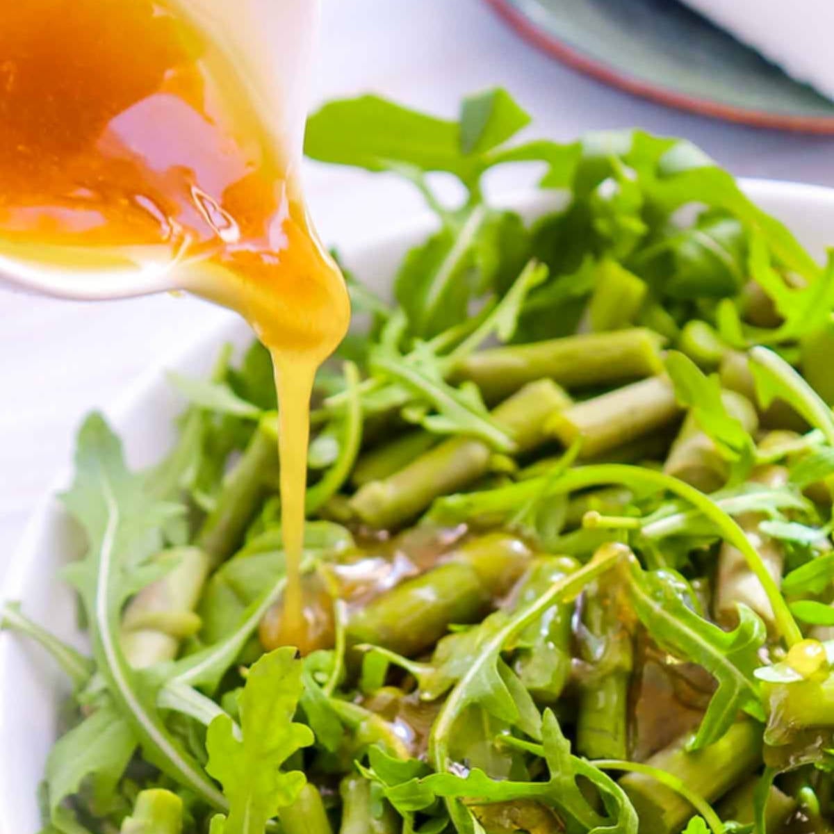 Honey vinaigrette being poured over asparagus and arugula in a white bowl