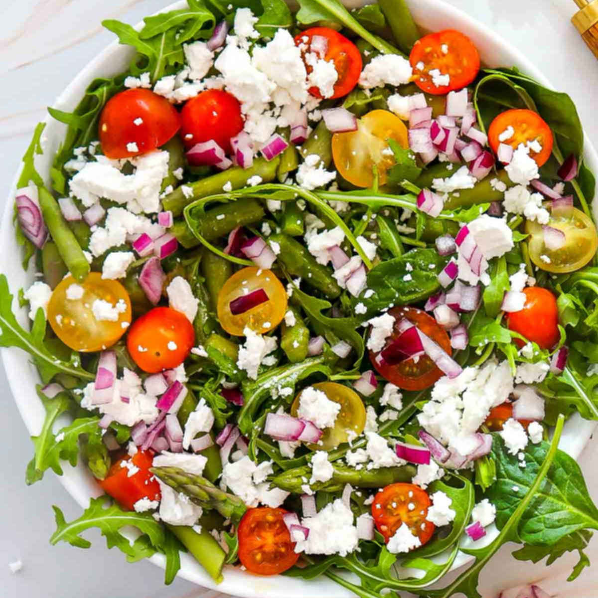 Finished goat cheese asparagus salad with arugula, cherry tomatoes, red onion, and crumbled goat cheese in a white bowl