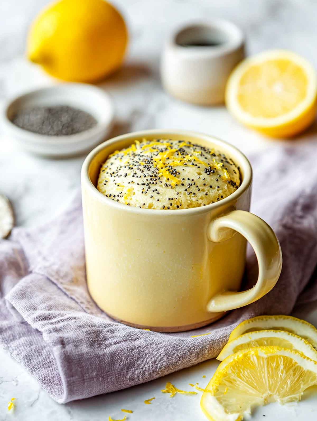 A bright angled view of a pale yellow ceramic mug filled with a lemon poppy seed mug cake, topped with fresh lemon zest and visible poppy seeds, set on a white marble countertop with lemon slices, a small bowl of Greek yogurt, and a soft lavender linen napkin nearby.